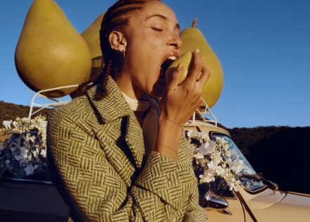 Adwoa Aboah, cheveux tressés, mordant dans une poire avec, en toile de fond, trois gros accessoires en forme de poire placés sur le toit d’une voiture.